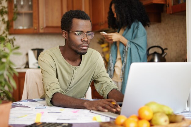 Junger afrikanischer Mann in den Gläsern, die vor offenem Laptop sitzen, sich auf Papierkram konzentrieren und inländische Rechnungen online bezahlen