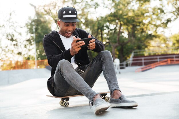 Junger afrikanischer Mann mit einem Skateboard unter Verwendung des Smartphones