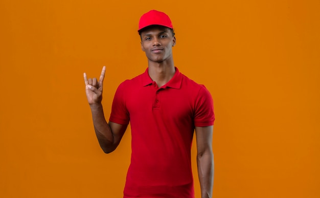 Kostenloses Foto junger afroamerikaner-auslieferungsmann, der rotes poloshirt und kappe trägt, die rockgeste macht, die über isoliertem orange steht