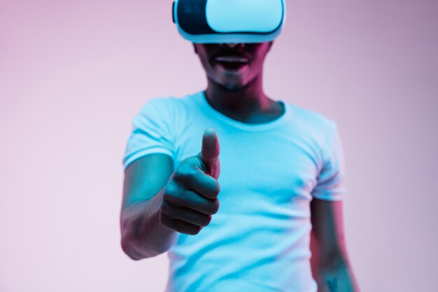 Junger Afroamerikaner in VR-Brille im Neonlicht auf Gradientenhintergrund. Männerporträt