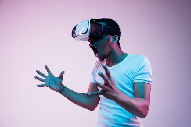 Junger Afroamerikaner spielt in VR-Brille im Neonlicht auf Gradientenhintergrund. Männerporträt