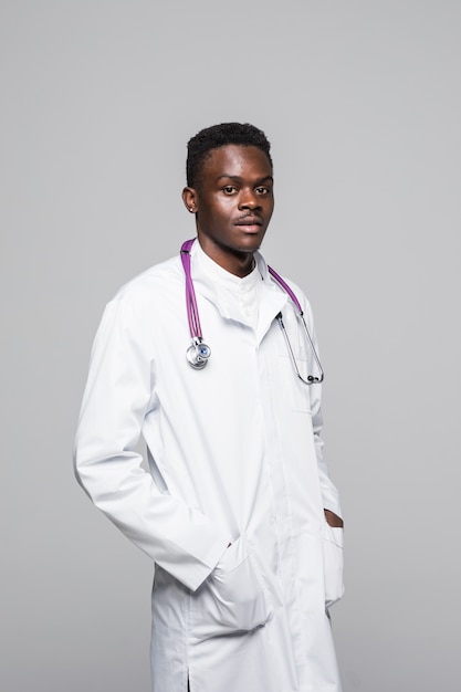 Junger afroamerikanischer Arzt in der weißen Uniform lokalisiert auf weißem Hintergrund, stehend mit Armen in der Tasche, die professionell und hochkompetent im Bereich der medizinischen Spezialisierung schaut