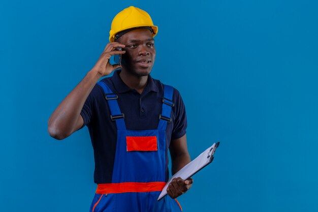 Junger afroamerikanischer Baumeister-Mann, der Bauuniform und Sicherheitshelm hält, der Klemmbrett hält, das auf Handy spricht, das auf Blau steht