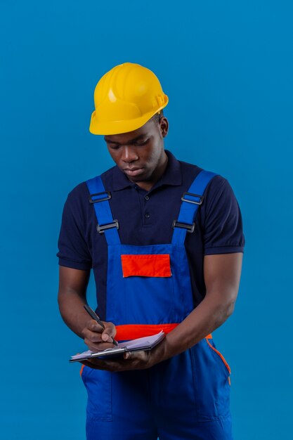 Junger afroamerikanischer Baumeistermann, der Bauuniform und Sicherheitshelm hält, die Klemmbrett hält, das Notizen mit Stift macht, der auf Blau steht