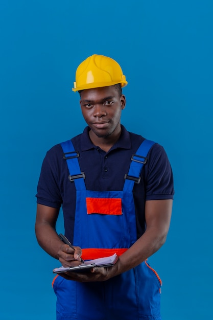 Junger afroamerikanischer Baumeistermann, der Bauuniform und Sicherheitshelm hält, die Klemmbrett und Stift hält, die Notizen mit freundlichem Lächeln machen, das auf Blau steht