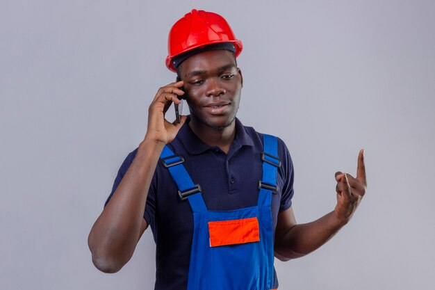 Junger afroamerikanischer Baumeistermann, der Bauuniform und Sicherheitshelm spricht, der auf Handy spricht, das Steinsymbol lächelnd stehend macht