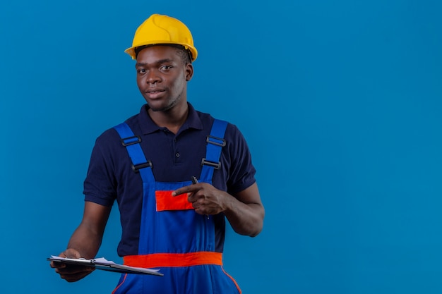 Junger afroamerikanischer Baumeistermann, der Bauuniform und Sicherheitshelm trägt, der mit Klemmbrett steht, das mit Zeigefinger mit zuversichtlichem Lächeln darauf zeigt