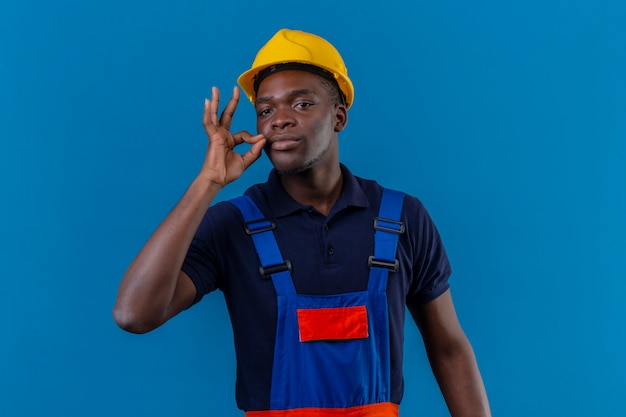 Junger afroamerikanischer Baumeistermann, der Bauuniform und Sicherheitshelm trägt, der Schweigengeste macht, wie das Schließen seines Mundes mit einem Reißverschluss auf lokalisiertem Blau tut