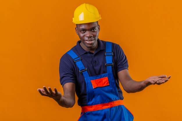 Junger afroamerikanischer Baumeistermann, der Bauuniform und Sicherheitshelm trägt, die mit aggressivem Ausdruck und Armen stehen stehen auf Orange stehen