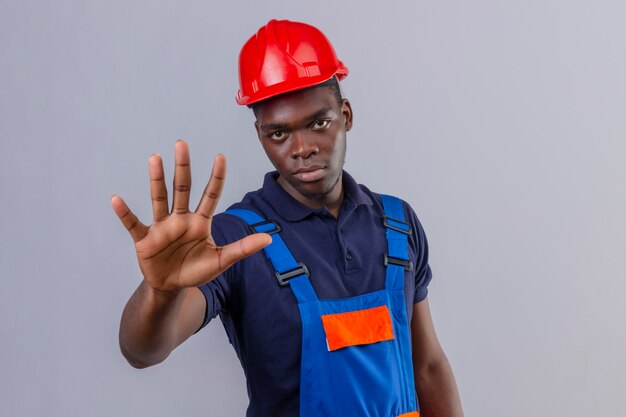 Junger afroamerikanischer Baumeistermann, der Bauuniform und Sicherheitshelm trägt, die mit offener Hand stehen Stoppschild mit ernsthafter und selbstbewusster Ausdrucksverteidigungsgeste stehen