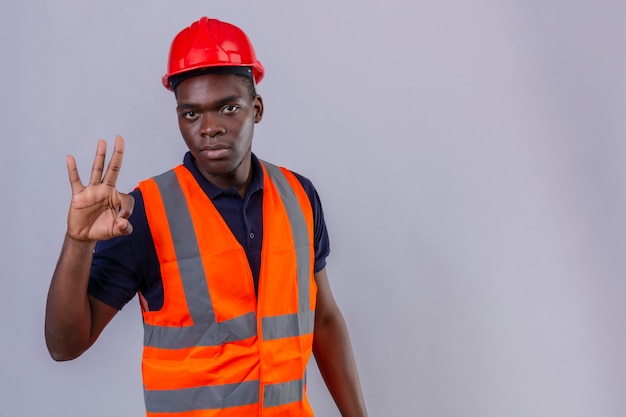 Junger afroamerikanischer Baumeistermann, der Bauweste und Sicherheitshelm mit ernstem Gesicht trägt, das ok Zeichen stehend tut