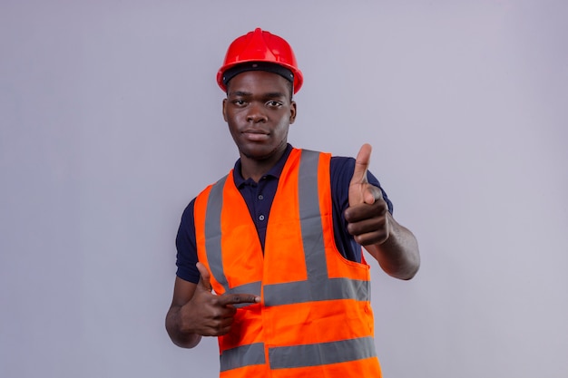 Junger afroamerikanischer Baumeistermann, der Bauweste und Sicherheitshelm trägt, der mit dem Finger zeigt, der selbstbewusst steht