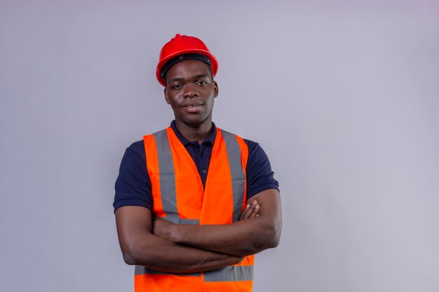 Junger afroamerikanischer Baumeistermann, der Bauweste und Sicherheitshelm trägt, die mit den Armen gekreuzt mit dem selbstbewussten Lächeln auf lokalisiertem Weiß stehen