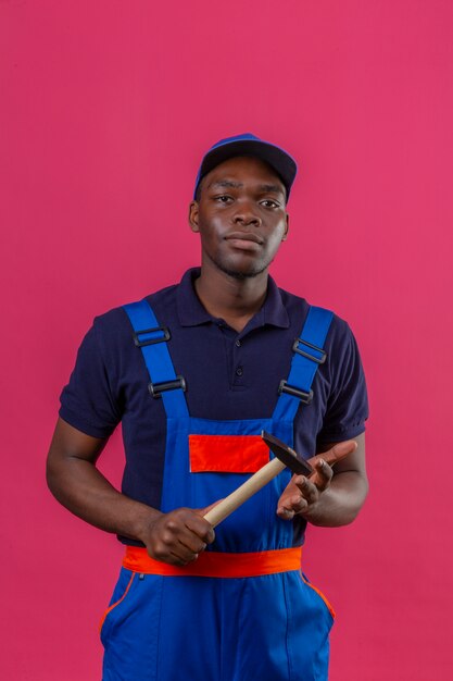 Junger afroamerikanischer Baumeistermann, der Konstruktionsuniform und Kappenhaltehammer in den Händen trägt, die zuversichtlich stehen auf Rosa stehen