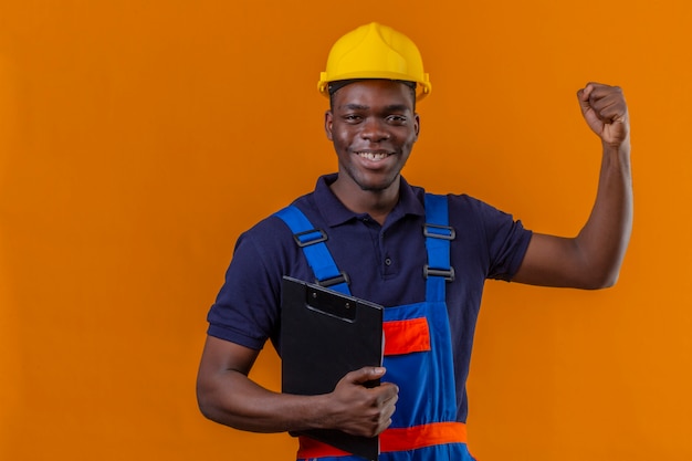 Junger afroamerikanischer Baumeistermann, der Konstruktionsuniform und Sicherheitshelm steht, der mit Klemmbrett anhebt, das Handballing-Faustlächeln stehend mit glücklichem Gesicht hebt, das Sieg feiert