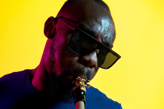 Junger afroamerikanischer Jazzmusiker, der das Saxophon auf gelbem Hintergrund im trendigen Neonlicht spielt.