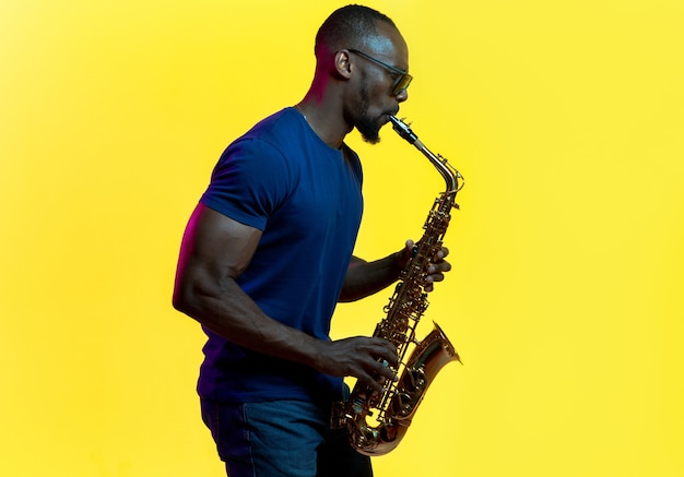 Junger afroamerikanischer jazzmusiker, der saxophon auf gelbem studiohintergrund in trendigem neonlicht spielt. konzept der musik, hobby. fröhlicher kerl improvisiert. buntes porträt des künstlers.