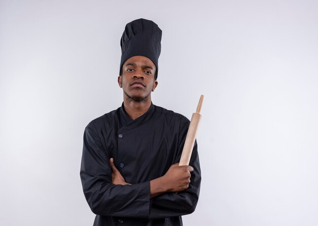 Junger afroamerikanischer Koch in der Kochuniform kreuzt Arme und hält Nudelholz lokalisiert auf weißem Hintergrund mit Kopienraum