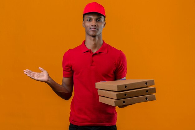 Junger afroamerikanischer Lieferbote, der rotes Poloshirt und Kappe hält, die Stapel von Pizzaschachteln hält, die verwirrt schauen und Zweifel über isoliertem Orange haben