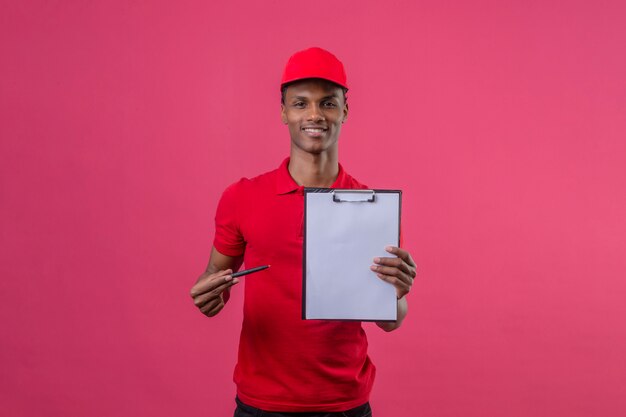 Junger afroamerikanischer Lieferbote, der rotes Poloshirt und Kappe hält, die Zwischenablage zeigt, die zur Kamera zeigt und mit Stift darauf zeigt, der über isoliertes Rosa lächelt