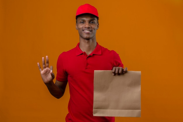 Junger afroamerikanischer Lieferbote, der rotes Poloshirt und Kappe mit Papiertüte mit Essen zum Mitnehmen trägt, das ok Zeichen oder Nummer drei mit Fingern über isoliertem Orange macht