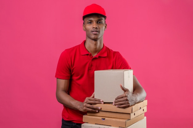 Junger afroamerikanischer Lieferbote, der rotes Poloshirt und Kappe mit Stapel von Kisten trägt, die Kamera über lokalisiertem Rosa betrachten