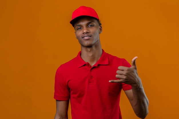 Junger afroamerikanischer Lieferbote, der rotes Poloshirt und Kappe trägt, die zuversichtlich macht, Telefongeste zu machen, rufen mich zurück Zeichen über isolierte Orange