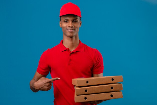 Junger afroamerikanischer Lieferbote, der rotes Poloshirt und Kappe zeigt, zeigt mit Finger zum Stapel von Pizzaschachteln in der anderen Hand über isoliertem Blau