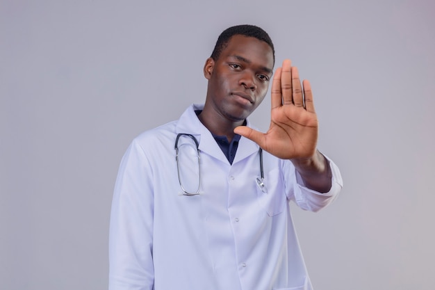 Junger afroamerikanischer männlicher Arzt, der weißen Kittel mit Stethoskop mit ernstem Gesicht mit offener Hand macht Stoppschild trägt
