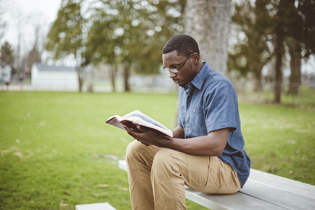 Junger afroamerikanischer Mann, der auf der Bank sitzt und die Bibel liest