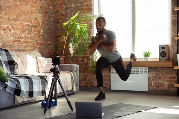 Junger afroamerikanischer Mann, der zu Hause Online-Kurse von Fitness, Aerobic, sportlichem Lebensstil während der Quarantäne, Neuaufnahme vor der Kamera, Streaming unterrichtet