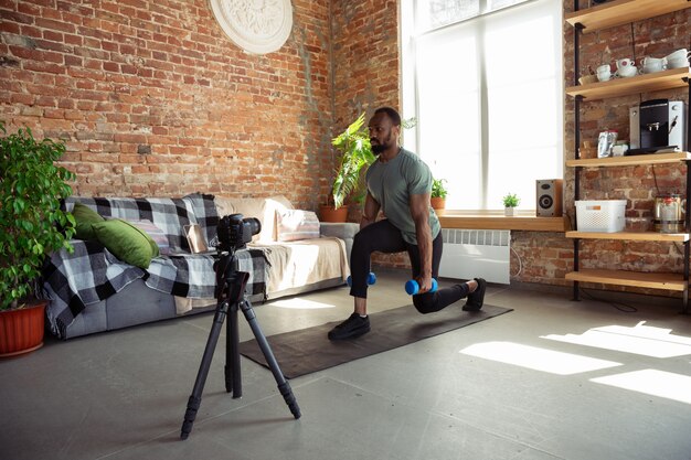 Junger afroamerikanischer Mann, der zu Hause Online-Kurse von Fitness, Aerobic, sportlichem Lebensstil während der Quarantäne, Neuaufnahme vor der Kamera, Streaming unterrichtet