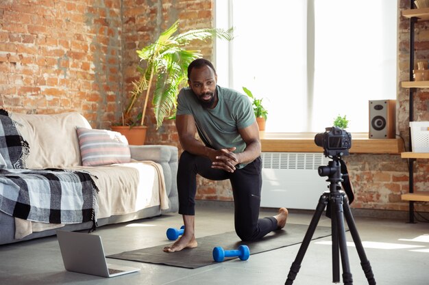 Junger afroamerikanischer Mann, der zu Hause Online-Kurse von Fitness, Aerobic, sportlichem Lebensstil während der Quarantäne, Neuaufnahme vor der Kamera, Streaming unterrichtet