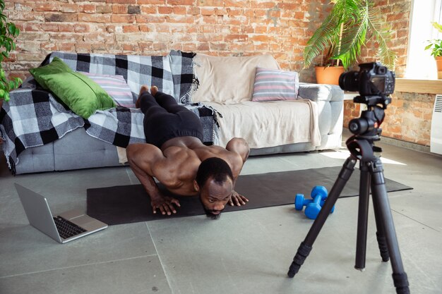 Junger afroamerikanischer Mann, der zu Hause Online-Kurse von Fitness, Aerobic, sportlichem Lebensstil während der Quarantäne, Neuaufnahme vor der Kamera, Streaming unterrichtet