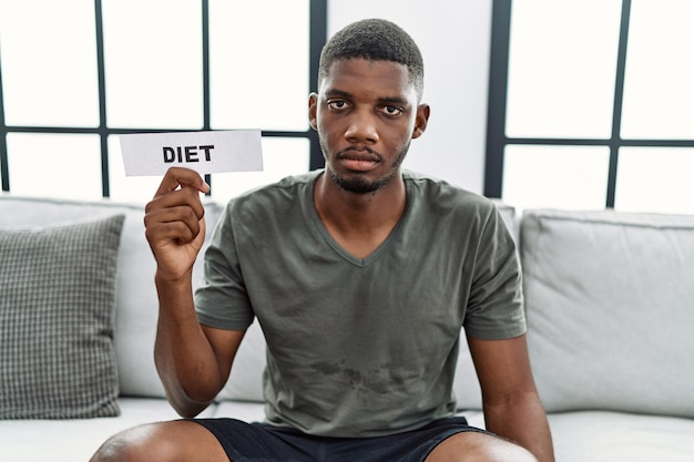 Junger afroamerikanischer Mann mit Diät-Banner sitzt zu Hause auf dem Sofa und denkt nachdenklich und nüchtern und sieht selbstbewusst aus