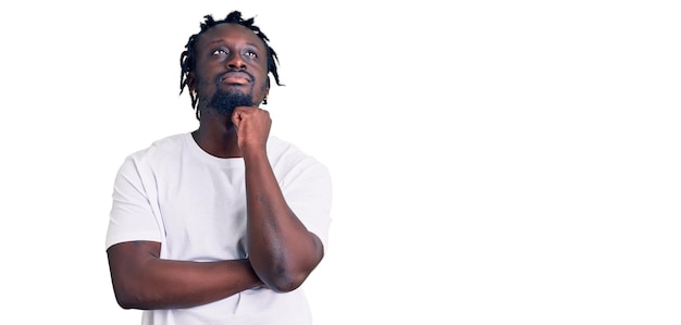 Junger afroamerikanischer mann mit zöpfen trägt ein lässiges weißes t-shirt mit der hand am kinn und denkt über fragen nach, nachdenklicher gesichtsausdruck, lächelndes und nachdenkliches gesichtszweifelkonzept