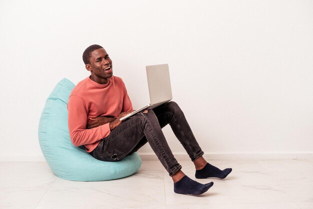 Junger afroamerikanischer mann sitzt auf einem puff mit laptop isoliert auf weißem hintergrund, lacht und hat spaß.