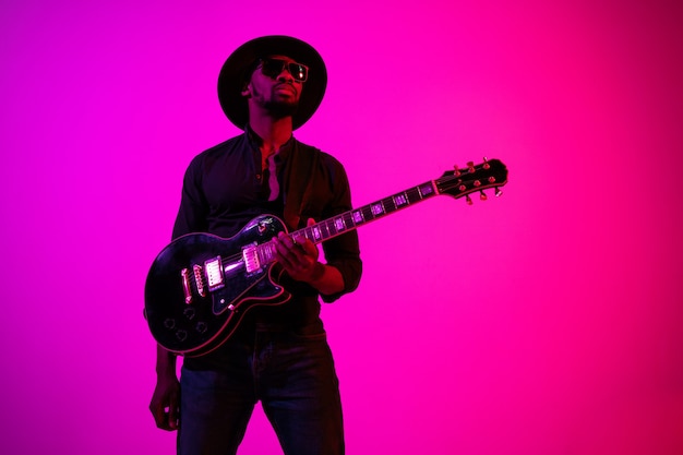 Junger afroamerikanischer musiker, der die gitarre wie ein rockstar auf lila-rosa hintergrund mit farbverlauf in neonlicht spielt.