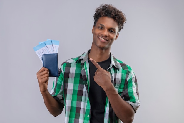 Junger afroamerikanischer Reisender Mann, der Flugtickets hält, die mit dem Finger auf sie zeigen, die fröhlich positiv und glücklich über weißem Hintergrund stehend lächeln