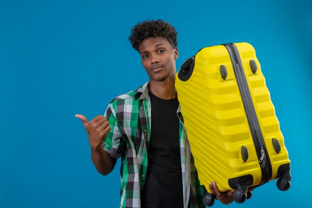 Junger afroamerikanischer Reisender Mann, der Koffer hält, der mit dem Finger zur Seite schaut, die Kamera mit dem zuversichtlichen Lächeln betrachtet, das über blauem Hintergrund steht