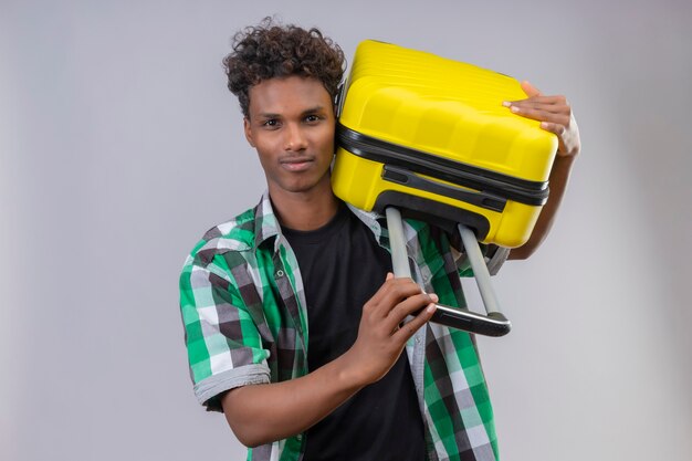 Junger afroamerikanischer Reisender Mann, der Koffer hält Kamera mit sicherem ernstem Ausdruck auf Gesicht, das über weißem Hintergrund steht