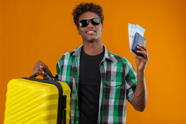 Junger afroamerikanischer Reisender Mann, der schwarze Sonnenbrille trägt, die mit Koffer hält, der Flugtickets hält, die fröhlich positiv und glücklich über orange Hintergrund lächeln