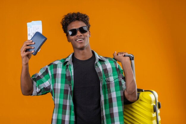 Junger afroamerikanischer Reisender Mann, der schwarze Sonnenbrille trägt, die mit Koffer hält, der Flugtickets hält, die fröhlich positiv und glücklich über orange Hintergrund lächeln