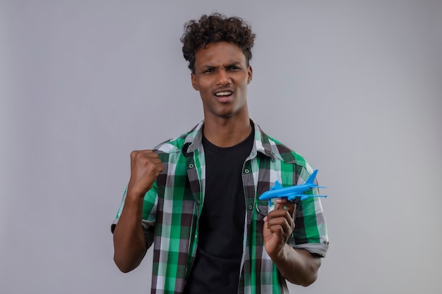 Junger afroamerikanischer Reisender Mann, der Spielzeugflugzeug hält, das Faust erhöht und freudiges Erhöhen der Faust freut sich über seinen Erfolg, der über weißem Hintergrund steht