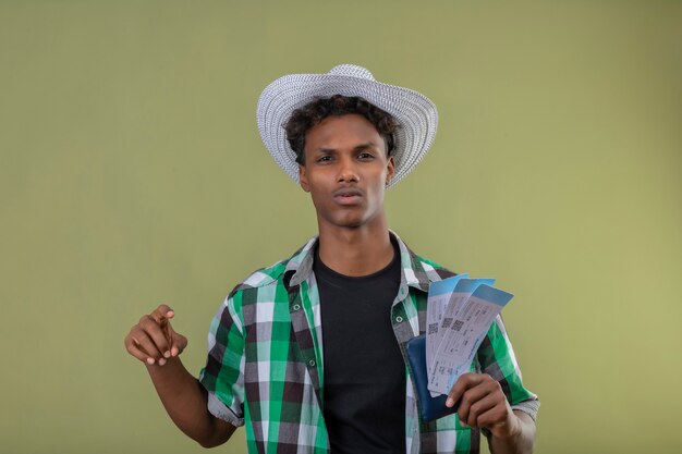 Junger afroamerikanischer Reisender Mann im Sommerhut, der Flugtickets hält Kamera mit ernstem zuversichtlichem Ausdruck betrachtet