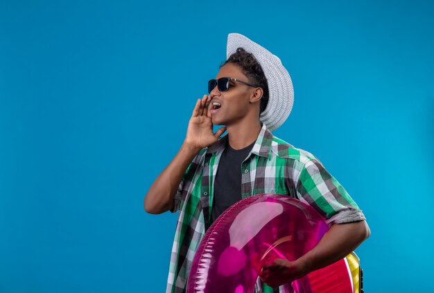 Junger afroamerikanischer Reisender Mann im Sommerhut, der schwarze Sonnenbrille hält, die aufblasbaren Ring hält, der beiseite schreit oder jemanden ruft, der über blauem Hintergrund steht