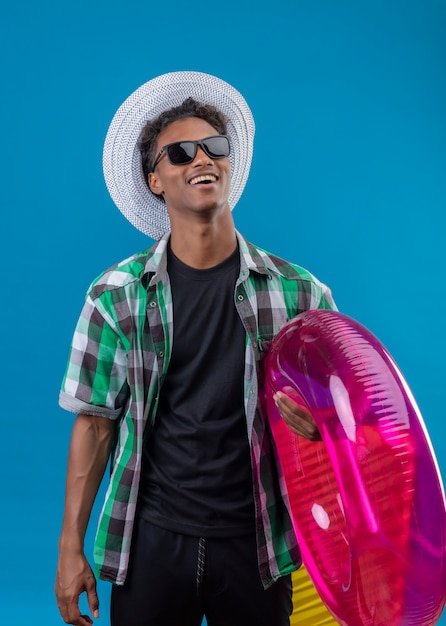 Junger afroamerikanischer Reisender Mann im Sommerhut, der schwarze Sonnenbrille hält, die aufblasbaren Ring hält, der fröhlich glücklich und positiv steht über blauem Hintergrund