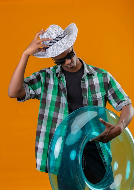 Junger afroamerikanischer Reisender Mann im Sommerhut, der schwarze Sonnenbrille hält, die aufblasbaren Ring hält, der unten schaut, der seinen Hut berührt