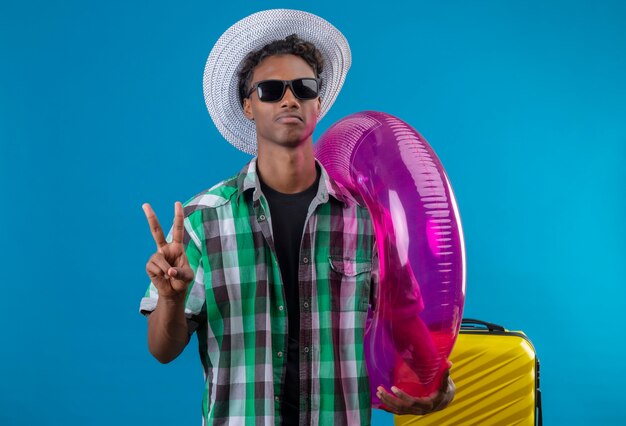 Junger afroamerikanischer Reisender Mann im Sommerhut, der schwarze Sonnenbrille mit Koffer hält aufblasbaren Ring, der Nummer zwei oder Siegeszeichen mit ernstem Gesicht zeigt