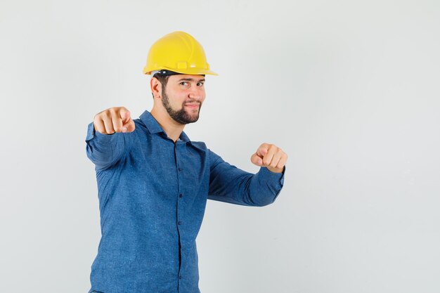 Junger Arbeiter, der auf Kamera in Hemd, Helm zeigt und optimistisch schaut.